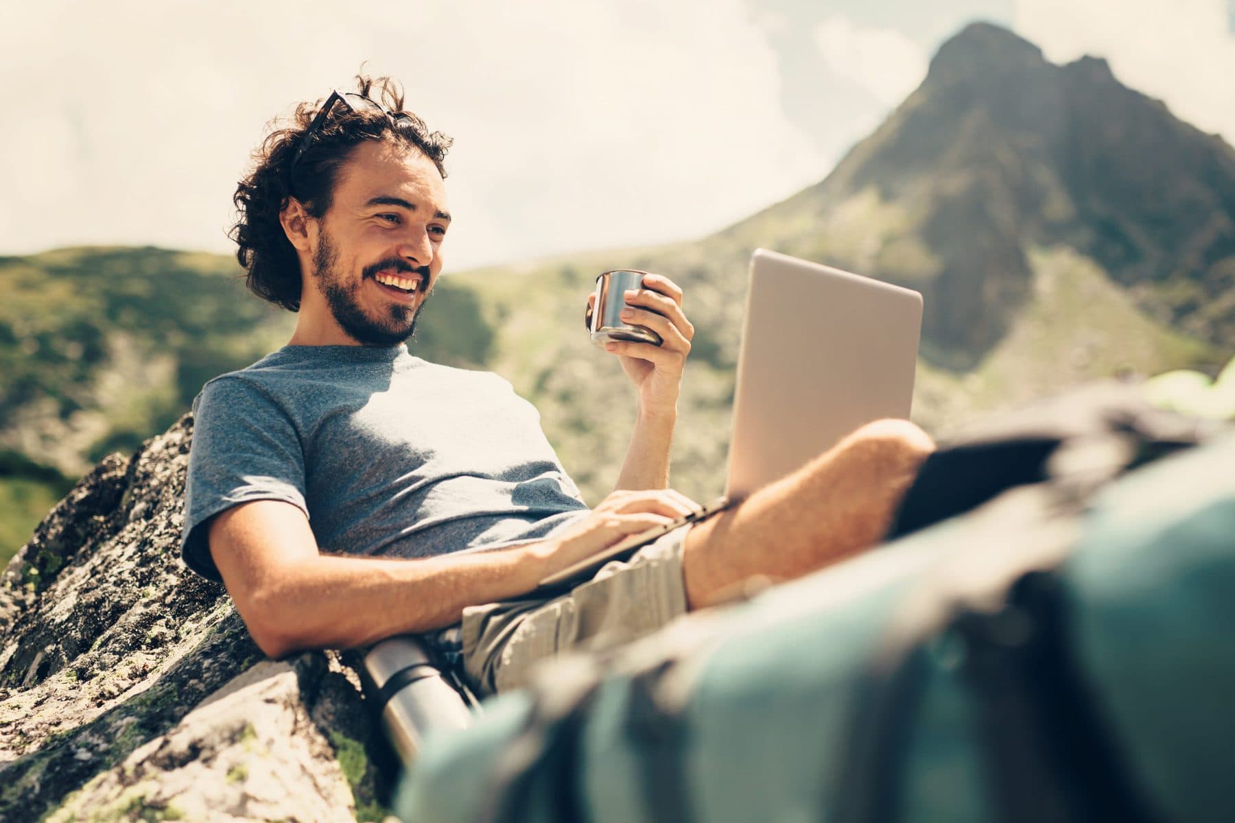 Millennial man enjoying a drink on the mountain while using a laptop